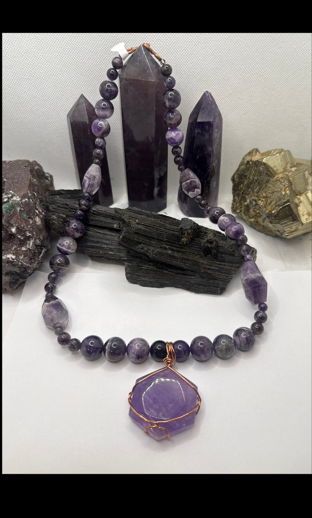 A purple amethyst gemstone beaded necklace with a faceted purple pendant wrapped in copper wire, displayed on a white surface with crystals and dark wood pieces in the background.