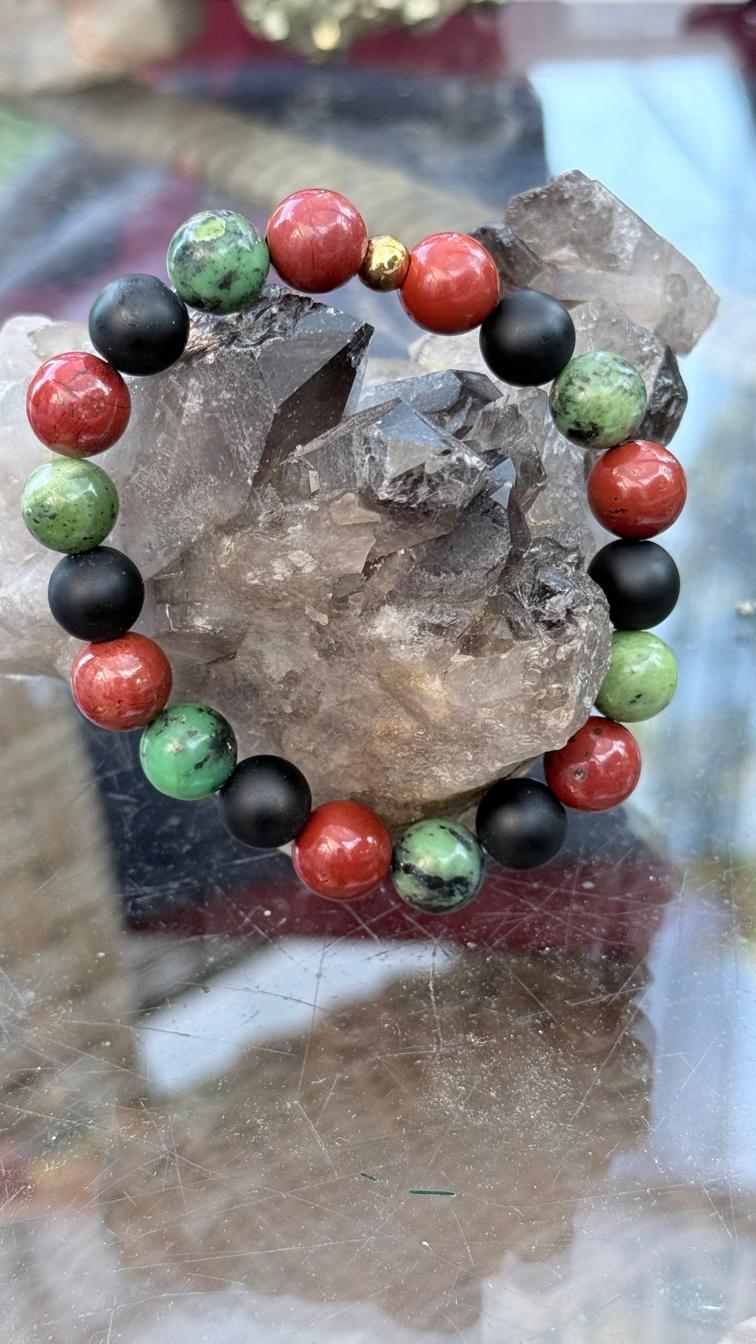 A beaded bracelet made of alternating red jasper, green chrysoprase and black onyx round beads,displayed on a clear quartz crystal cluster.