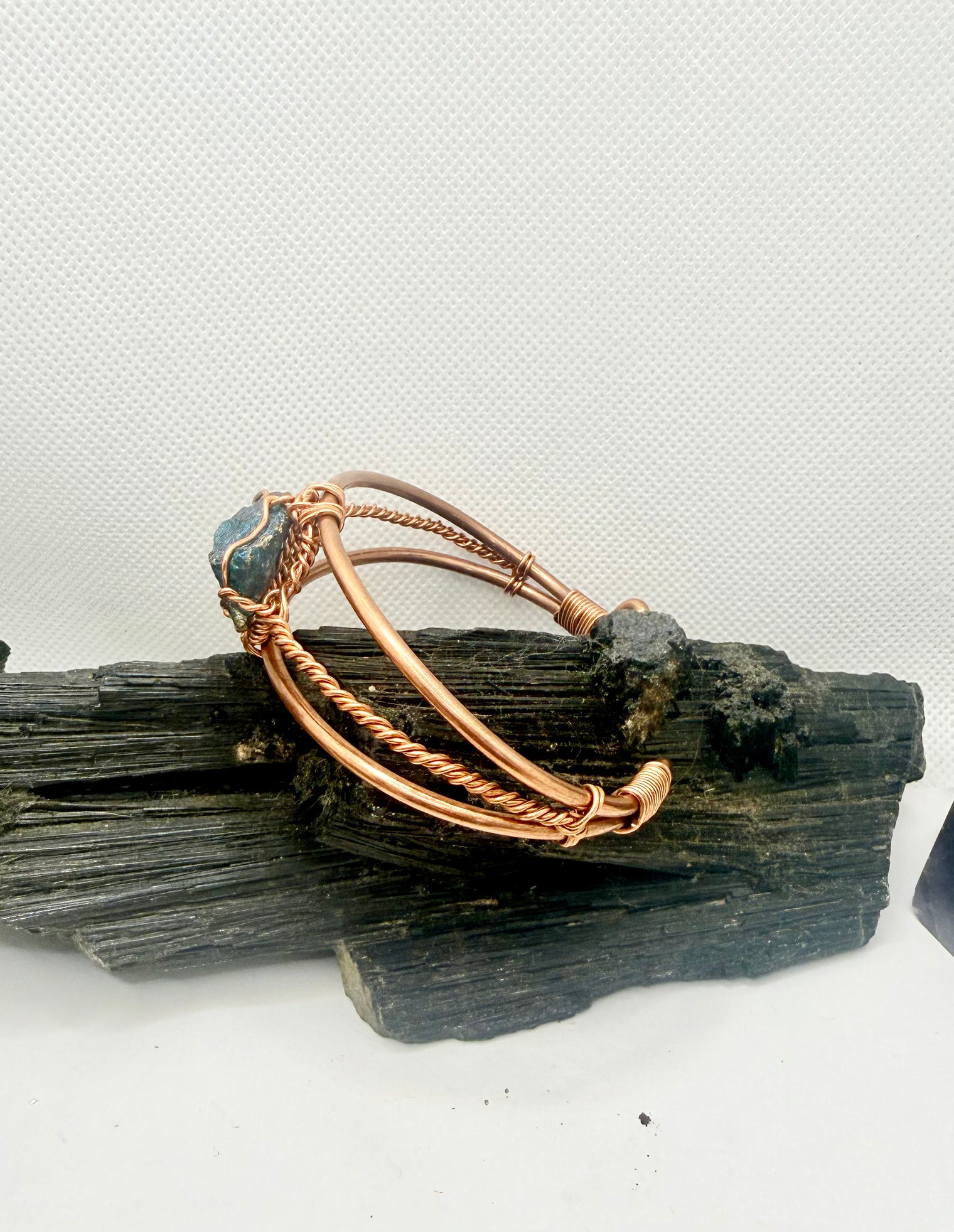 A copper-toned wire bangle bracelet with a small blue-green stone accent, displayed on a piece of dark wood against a light gray background.
chalcopyrite on copper wirewrapped bracelet on black tourmaline with white background