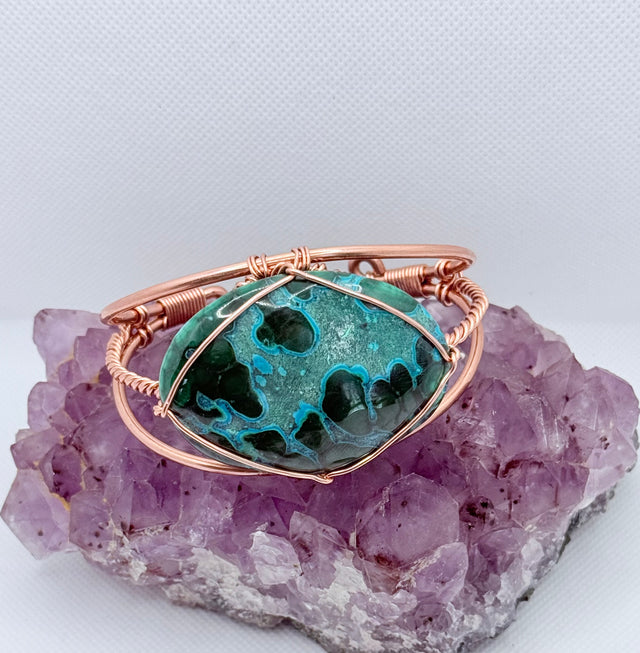 Jewelry with a green stone and rose gold wire wrapping on a purple crystal background the center stone of the copper bracelet is malachite chrysocolla