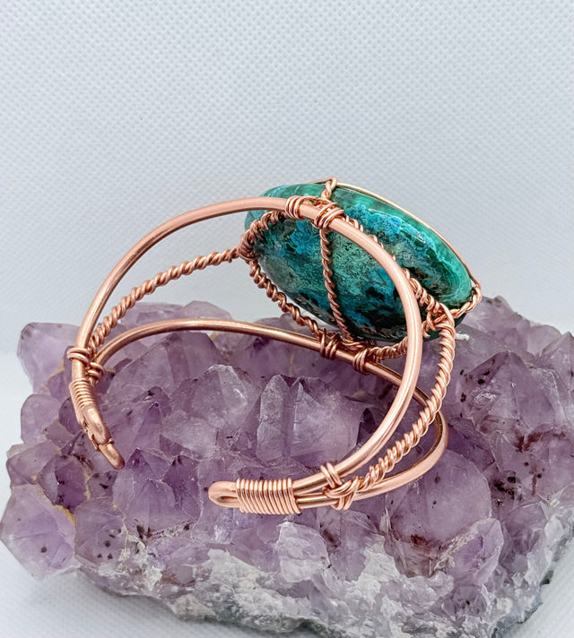 Jewelry with a green stone and rose gold wire wrapping on a purple crystal background the center stone of the copper bracelet is malachite chrysocolla