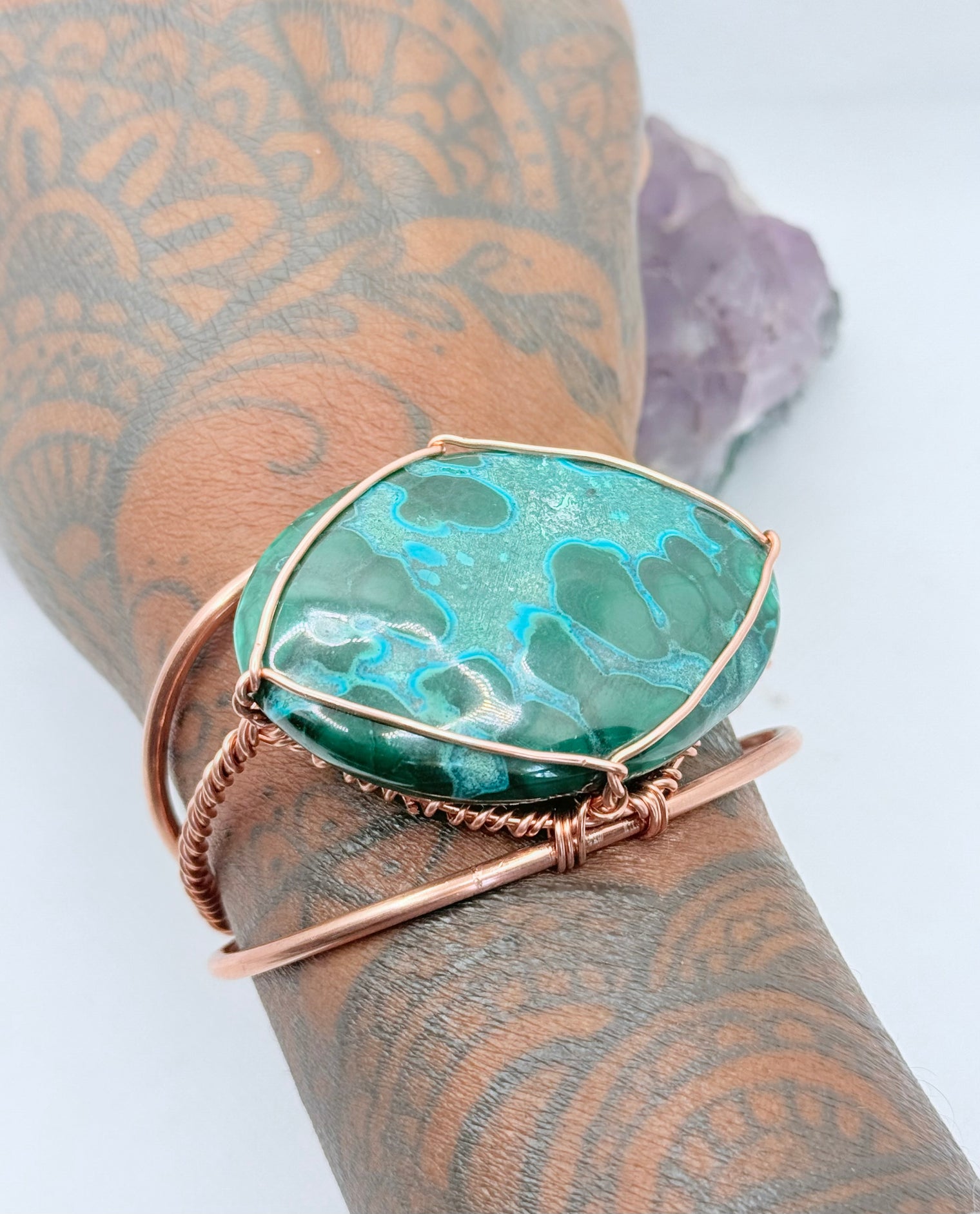 Jewelry with a green stone and rose gold wire wrapping on a purple crystal background the center stone of the copper bracelet is malachite chrysocolla
