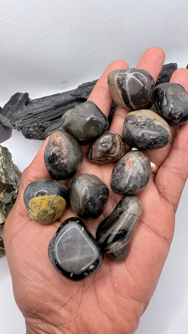 A hand holding a cluster of polished dark gray and black onyx tumbled stones, with rough mineral pieces in the background against a white backdrop.