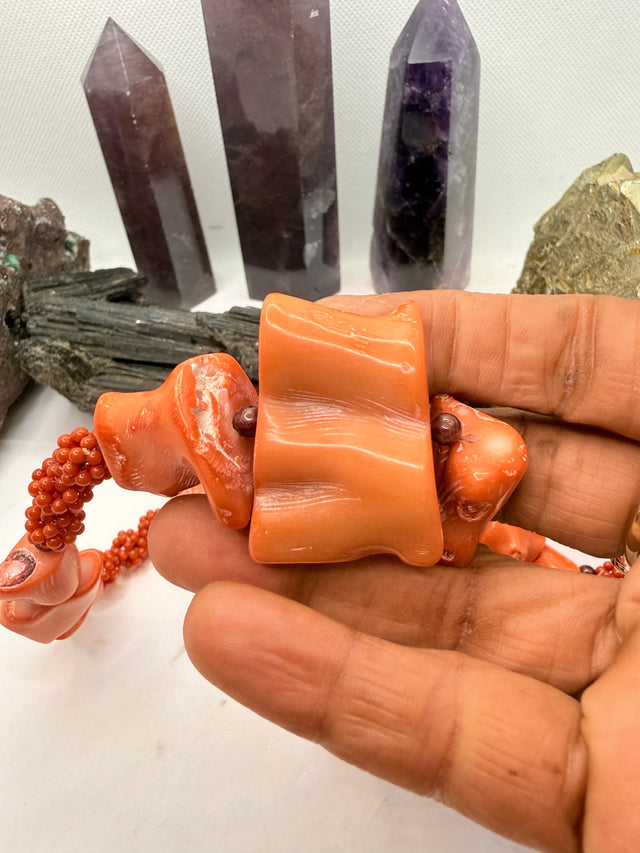 A pink and red coral coral bead necklace with chunky coral beads and small round garnet beads, displayed on a white surface with crystal points and rocks in the background.