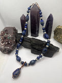 A beaded necklace made of dark blue lapis lazuli stones with silver spacer beads and a copper-wrapped teardrop pendant, displayed on a black wood block with purple crystal points and rocks in the background.