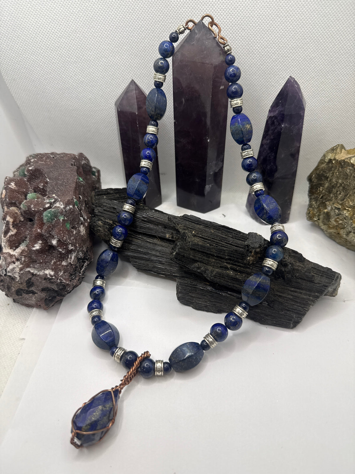 A beaded necklace made of dark blue lapis lazuli stones with silver spacer beads and a copper-wrapped teardrop pendant, displayed on a black wood block with purple crystal points and rocks in the background.