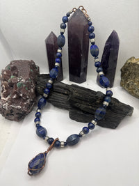 A beaded necklace made of dark blue lapis lazuli stones with silver spacer beads and a copper-wrapped teardrop pendant, displayed on a black wood block with purple crystal points and rocks in the background.
