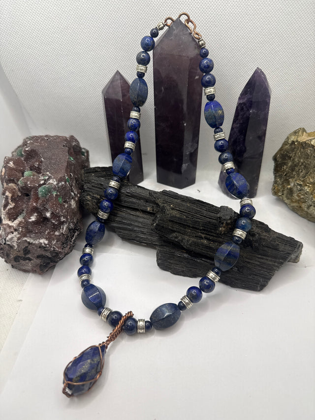 A beaded necklace made of dark blue lapis lazuli stones with silver spacer beads and a copper-wrapped teardrop pendant, displayed on a black wood block with purple crystal points and rocks in the background.