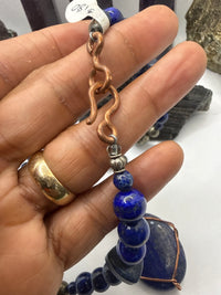 A close-up of a hand holding a beaded necklace featuring deep blue lapis lazuli stones, a copper-toned wavy chain, and silver spacer beads.