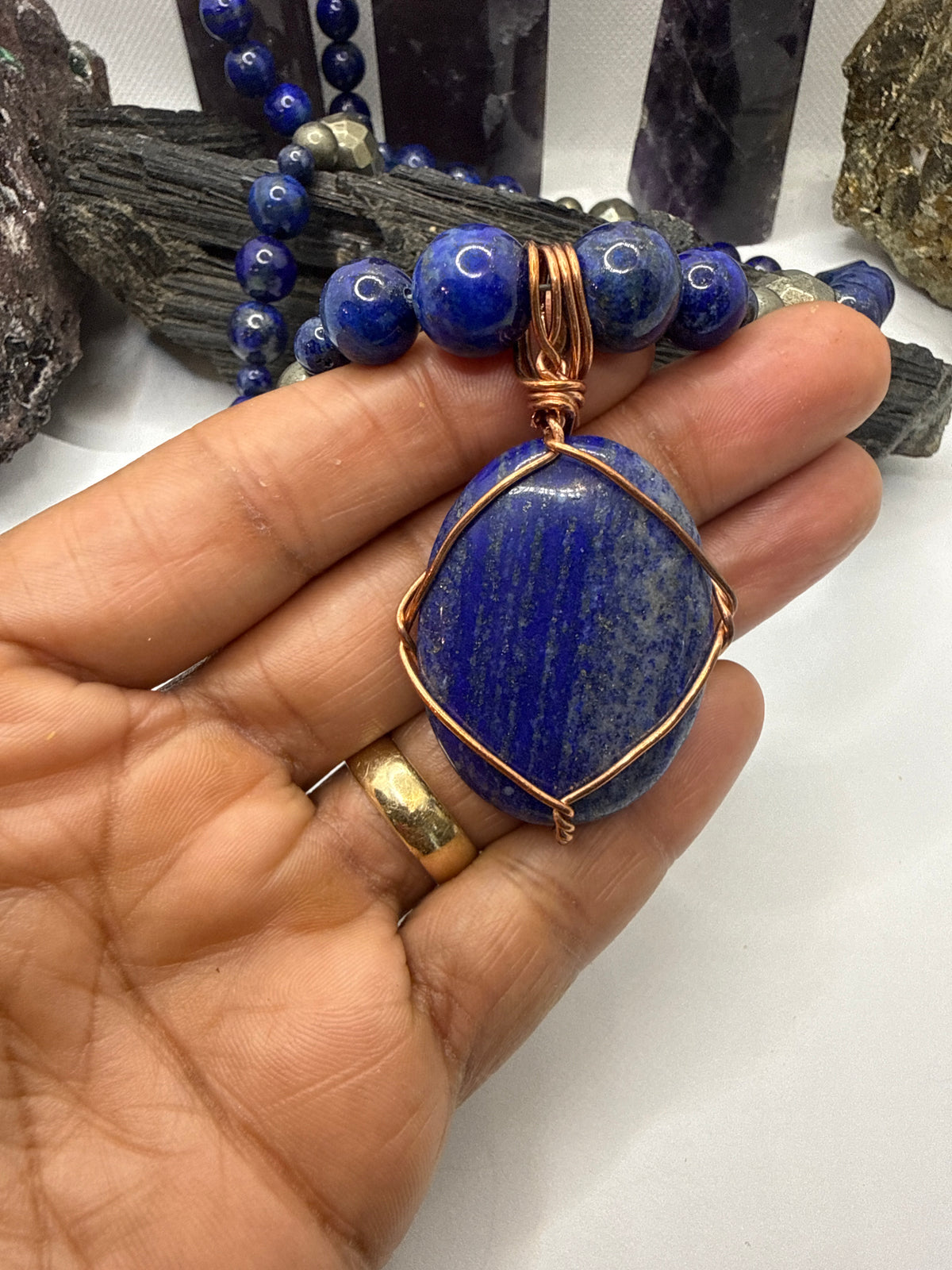 A close-up of a hand holding a blue lapis lazuli pendant with copper wire wrapping, with matching blue beads and crystals in the background.