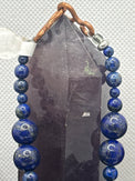 A beaded jewelry piece featuring deep blue lapis lazuli sphere beads draped over a dark gray crystal point, secured with a copper-colored metal clasp on a textured white background.