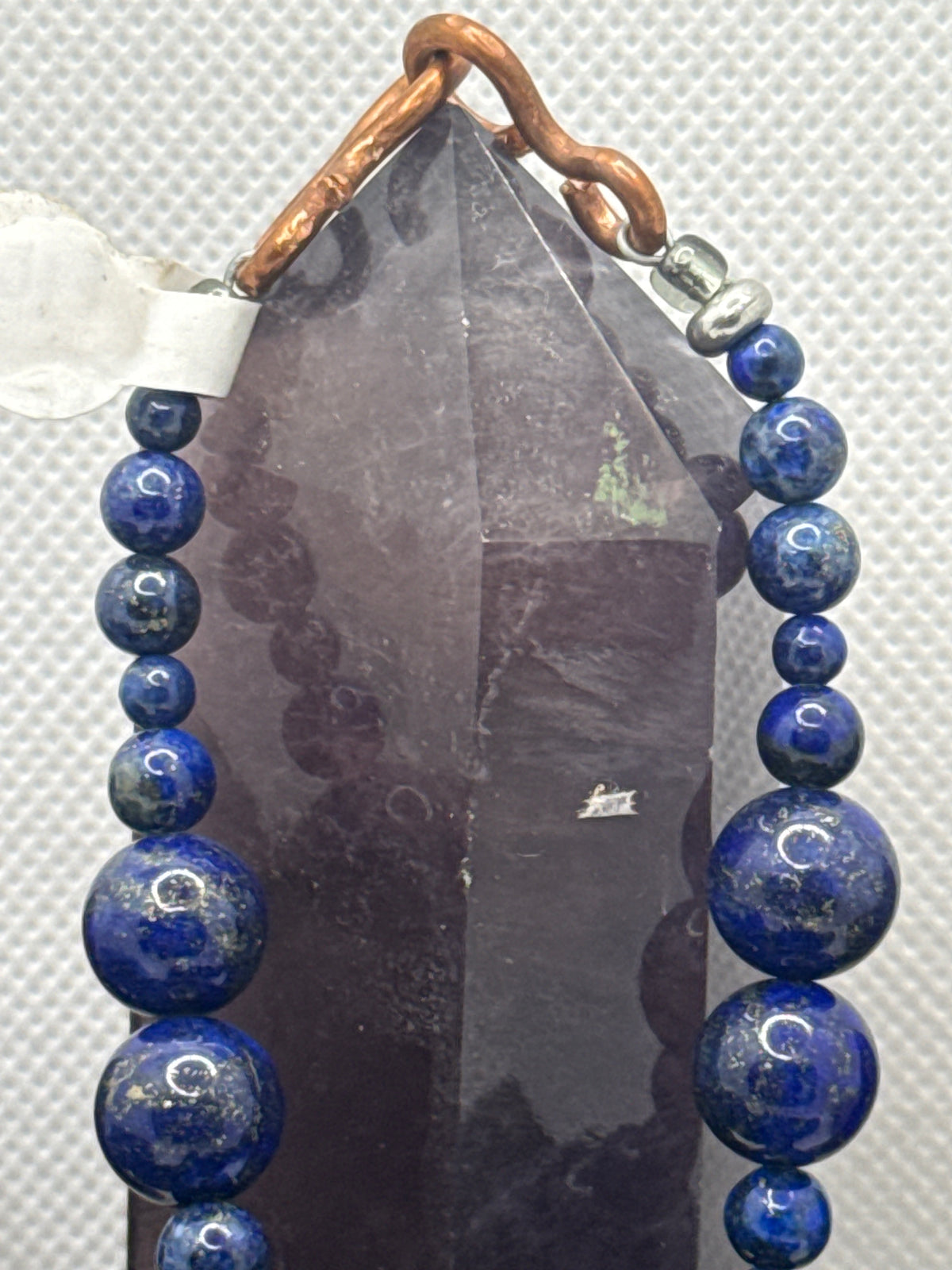 A beaded jewelry piece featuring deep blue lapis lazuli sphere beads draped over a dark gray crystal point, secured with a copper-colored metal clasp on a textured white background.