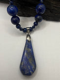 A lapis lazuli necklace featuring a large teardrop-shaped blue stone pendant with gold veining, suspended from a beaded strand of matching blue beads with silver-toned metal accents, displayed against a white background with a dark wood prop.