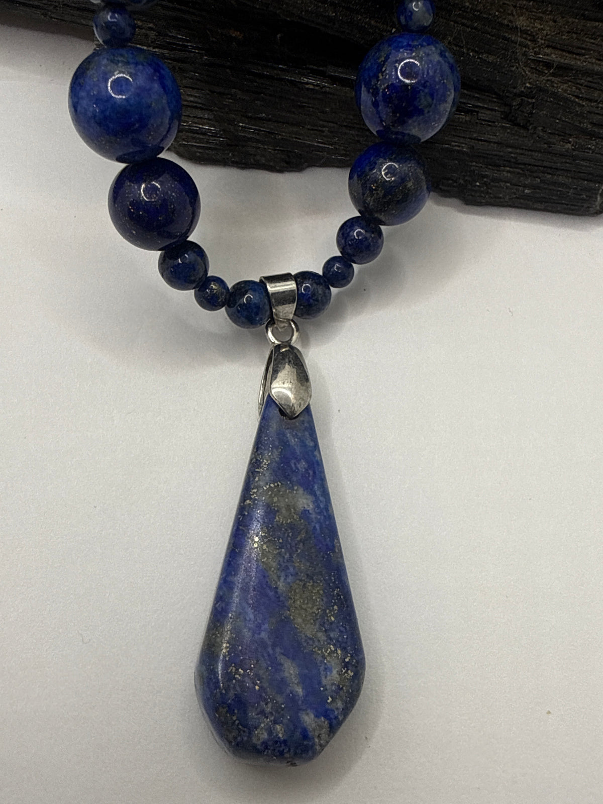A lapis lazuli necklace featuring a large teardrop-shaped blue stone pendant with gold veining, suspended from a beaded strand of matching blue beads with silver-toned metal accents, displayed against a white background with a dark wood prop.