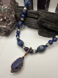 A beaded necklace with dark blue lapis lazuli stones, silver spacer beads, and a large teardrop-shaped lapis lazuli pendant wrapped in copper wire, displayed on a white surface with dark wood and crystals.