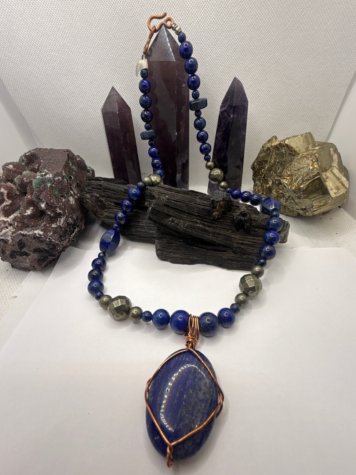 A beaded necklace with deep blue lapis lazuli-style beads, gray pyrite beads, and a large oval blue stone pendant wrapped in copper wire, displayed on a white surface with crystal points and rocks in the background.