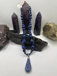 A beaded necklace with deep blue lapis lazuli stones, featuring a large teardrop-shaped blue stone pendant, displayed on a piece of dark wood with purple and gold crystals in the background.