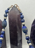 A beaded necklace made of deep blue lapis lazuli stones with silver-toned spacer beads and a gold-toned clasp, displayed against a dark crystal point on a light textured background.