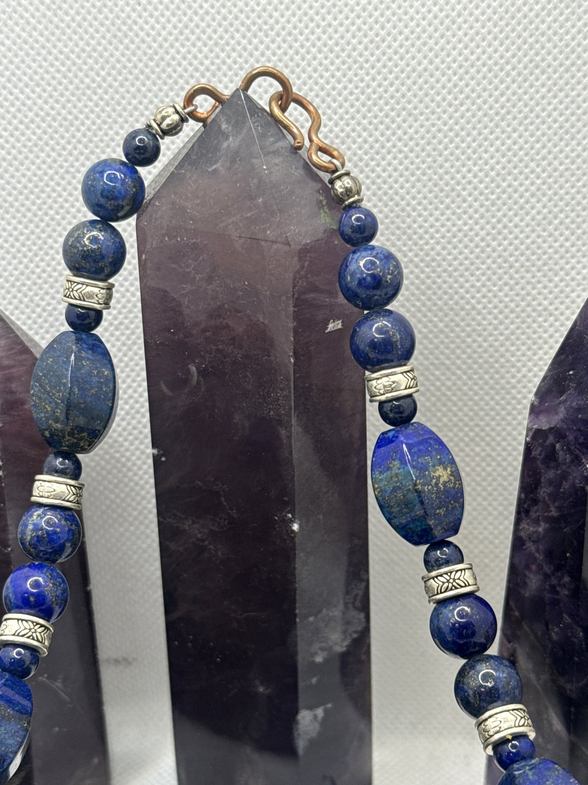 A beaded necklace made of deep blue lapis lazuli stones with silver-toned spacer beads and a gold-toned clasp, displayed against a dark crystal point on a light textured background.