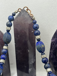A beaded necklace made of deep blue lapis lazuli stones with silver-toned spacer beads and a gold-toned clasp, displayed against a dark crystal point on a light textured background.