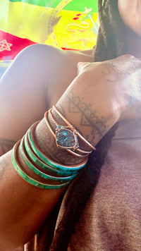 A close-up of a person's tattooed wrist wearing multiple bracelets, including a rose gold bangle with a blue-green stone centerpiece and stacked turquoise green bangles, against a warm-toned background. chalcopyrite on copper wirewrapped bracelet on black tourmaline with white background
