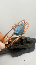 A handmade copper wire bangle bracelet featuring a raw, textured blue opal stone, held by a hand over a piece of dark wood against a white textured background.
chalcopyrite on copper wirewrapped bracelet on black tourmaline with white background
