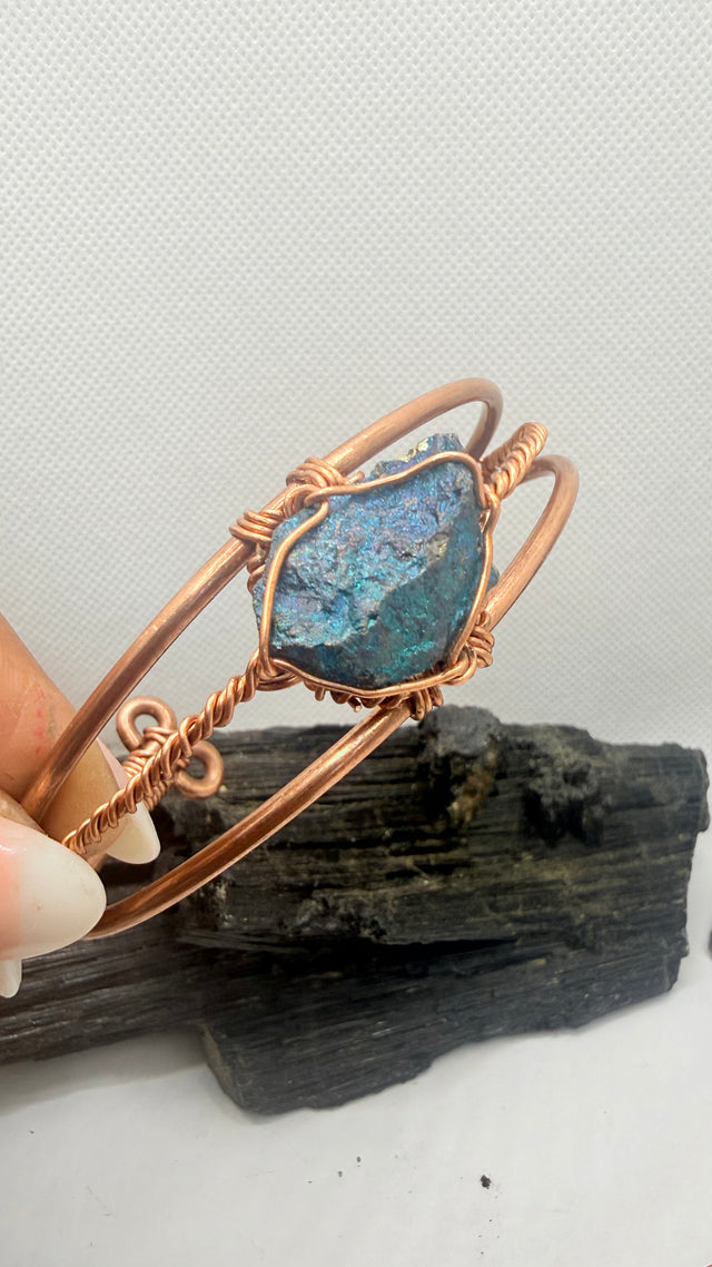 A handmade copper wire bangle bracelet featuring a raw, textured blue opal stone, held by a hand over a piece of dark wood against a white textured background.
chalcopyrite on copper wirewrapped bracelet on black tourmaline with white background
