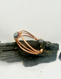 A copper-toned wire bangle bracelet with a small blue-green stone accent, displayed on a piece of dark wood against a light gray background.
chalcopyrite on copper wirewrapped bracelet on black tourmaline with white background
