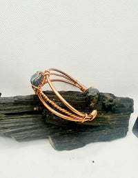 A copper-toned wire bangle bracelet with a small blue-green stone accent, displayed on a piece of dark wood against a light gray background.
chalcopyrite on copper wirewrapped bracelet on black tourmaline with white background
