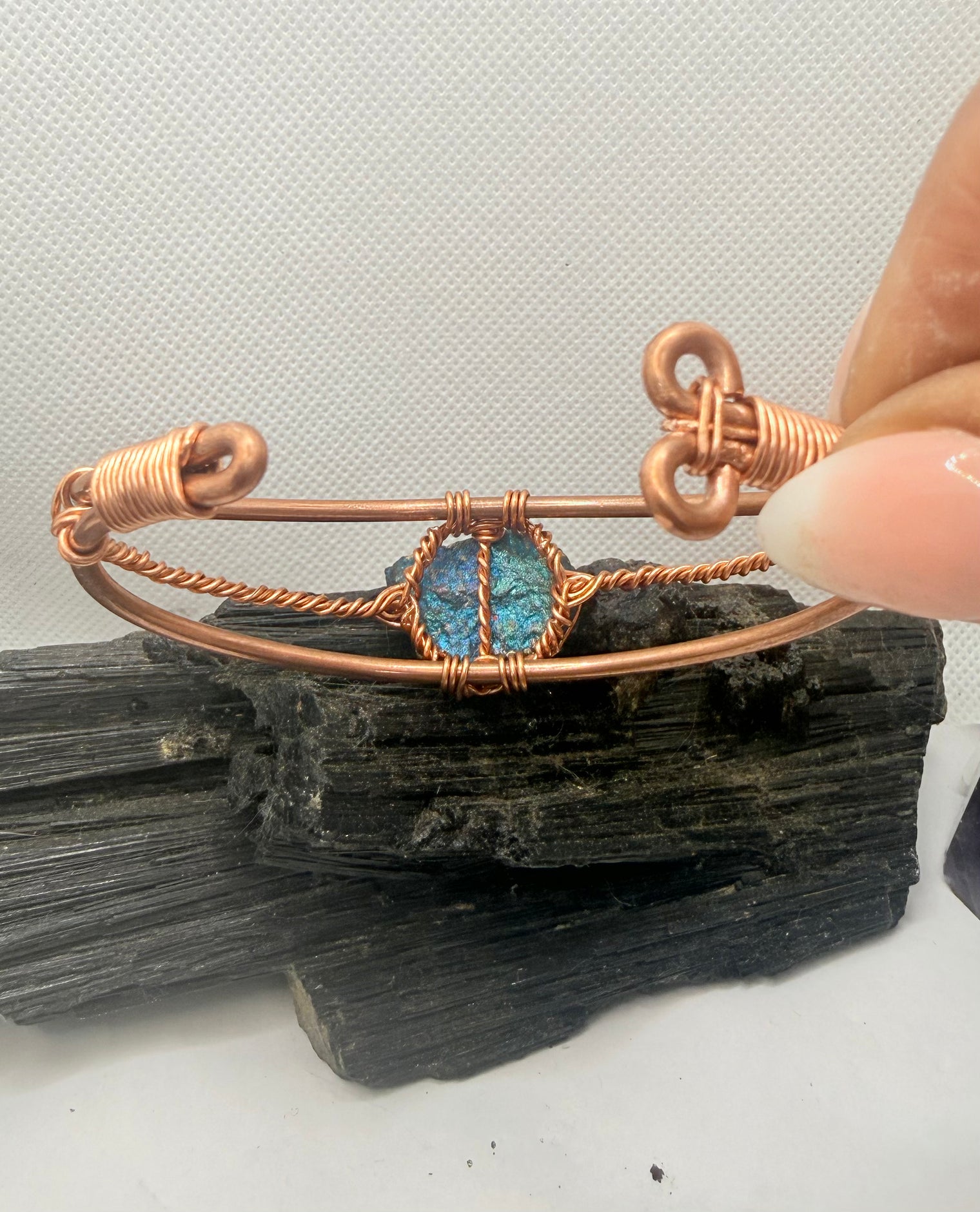 A copper wire cuff bracelet with a central blue-green opal-like stone, held by a hand over a dark mineral specimen on a white background.chalcopyrite on copper wirewrapped bracelet on black tourmaline with white background

