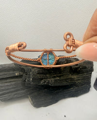 A copper wire cuff bracelet with a central blue-green opal-like stone, held by a hand over a dark mineral specimen on a white background.chalcopyrite on copper wirewrapped bracelet on black tourmaline with white background
