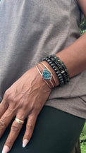 A close-up of a person's hand and wrist wearing multiple bracelets, including a copper-toned wrap bracelet with a blue-green stone and dark beaded bracelets, against a gray top. chalcopyrite on copper wirewrapped bracelet on black tourmaline with white background
