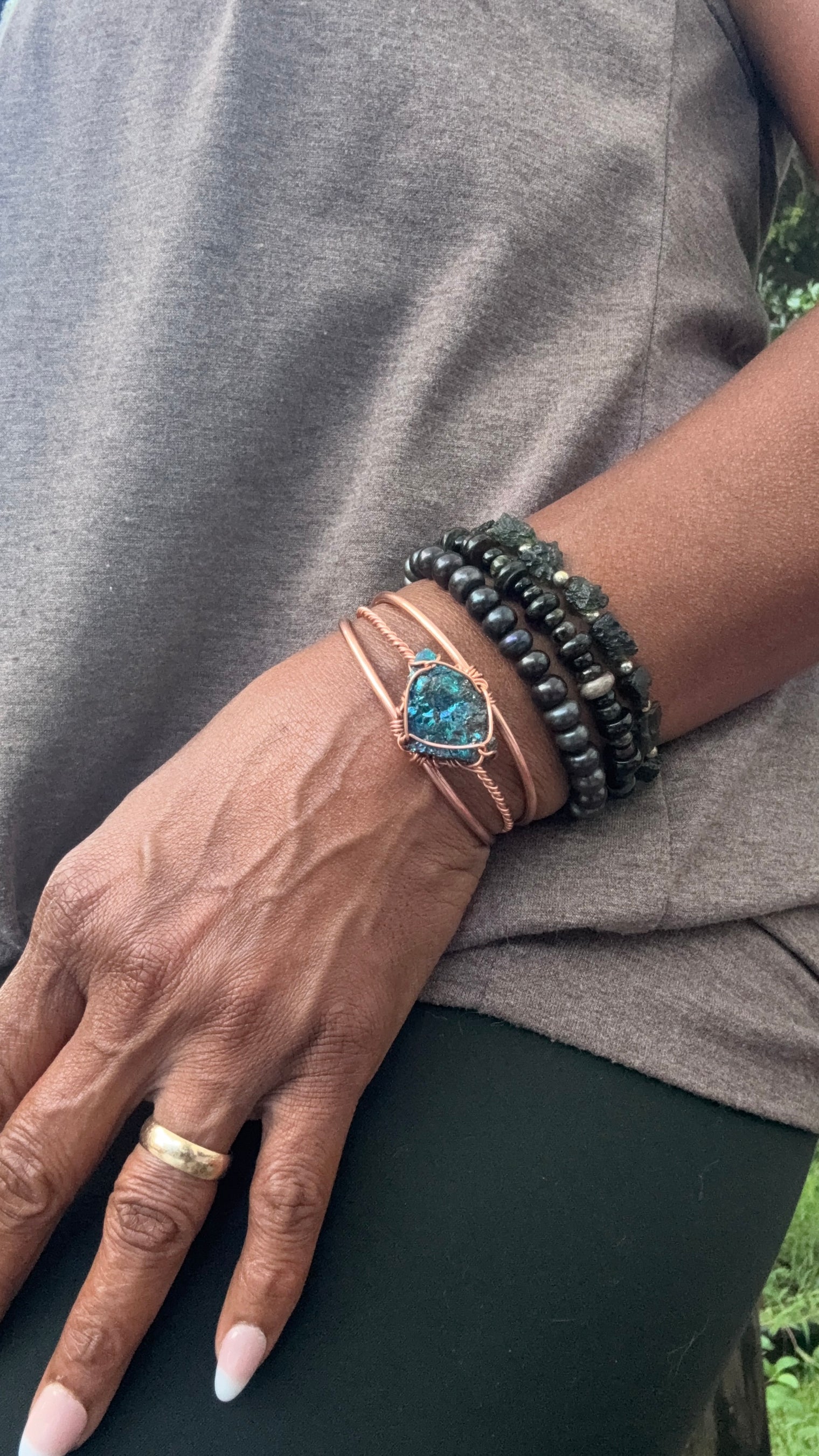 A close-up of a person's hand and wrist wearing multiple bracelets, including a copper-toned wrap bracelet with a blue-green stone and dark beaded bracelets, against a gray top. chalcopyrite on copper wirewrapped bracelet on black tourmaline with white background
