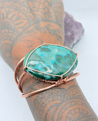 Jewelry with a green stone and rose gold wire wrapping on a purple crystal background the center stone of the copper bracelet is malachite chrysocolla 