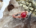 A handmade copper wire ring featuring a polished round red jade gemstone, displayed on a cluster of smoky quartz crystals with metallic mineral specimens in the background.