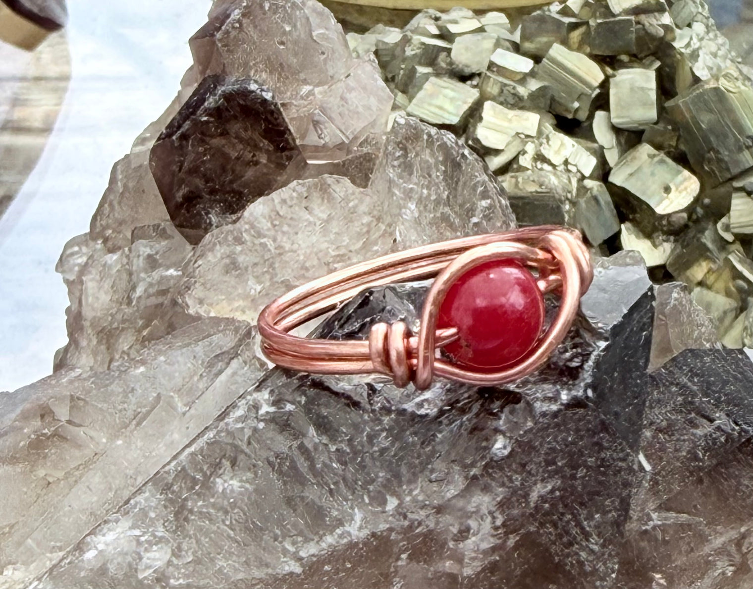 A handmade copper wire ring featuring a polished round red jade gemstone, displayed on a cluster of smoky quartz crystals with metallic mineral specimens in the background.