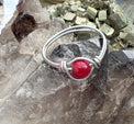 A handmade silver wire ring featuring a round, polished red jade gemstone, displayed on a textured gray crystal cluster.