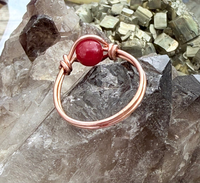A handmade ring featuring a round red jade 
gemstone wrapped in copper-colored wire, displayed on a textured gray crystal cluster.