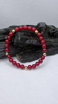 A beaded stretch bracelet made of round dark red jade beads with small gold spacer beads, displayed on a piece of dark wood against a light gray background.