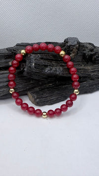 A beaded stretch bracelet made of deep red jade round beads with small gold spacer beads, displayed on a dark piece of wood against a light gray background.