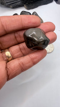 A hand holds a polished dark gray black onyx tumbled stone, with several other dark stones and a coin in the background on a white surface.