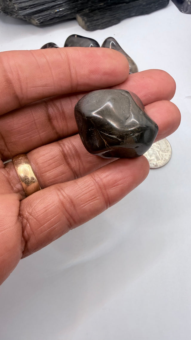 A hand holds a polished dark gray black onyx tumbled stone, with several other dark stones and a coin in the background on a white surface.
