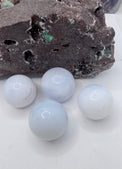 Four smooth, pale blue-white chalcedony spherical stones arranged on a white surface in front of a dark textured rock.