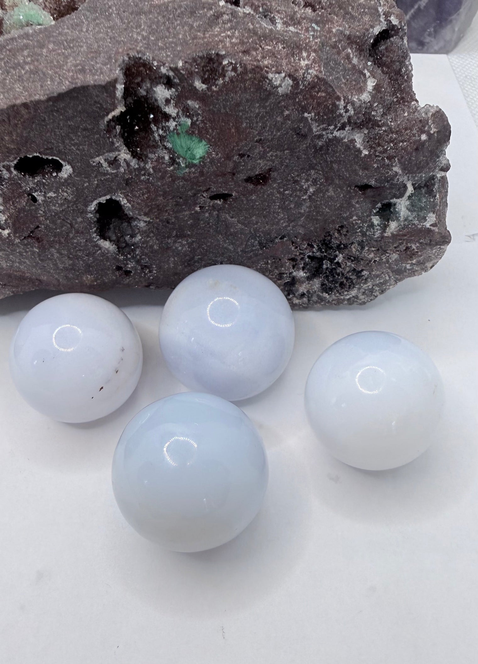 Four smooth, pale blue-white chalcedony spherical stones arranged on a white surface in front of a dark textured rock.