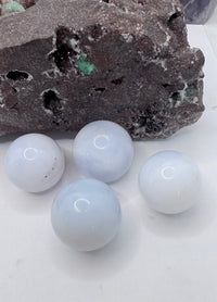 Four smooth, pale blue-white chalcedony spherical stones arranged on a white surface in front of a dark textured rock.