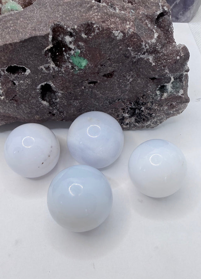 Four smooth, pale blue-white chalcedony spherical stones arranged on a white surface in front of a dark textured rock.