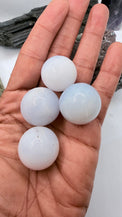 Four smooth, round, milky white blue chalcedony gemstone balls held in a person's open hand, with a dark mineral specimen visible in the background.
