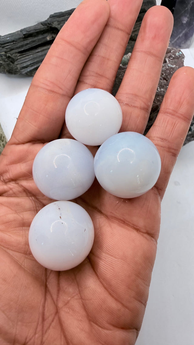 Four smooth, round, milky white blue chalcedony gemstone balls held in a person's open hand, with a dark mineral specimen visible in the background.