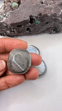 A hand holds a polished gray blue indian opal evil eye 
tumbled stone with a heart-shaped cutout, with two more polished stones and a rough stone in the background on a white surface.