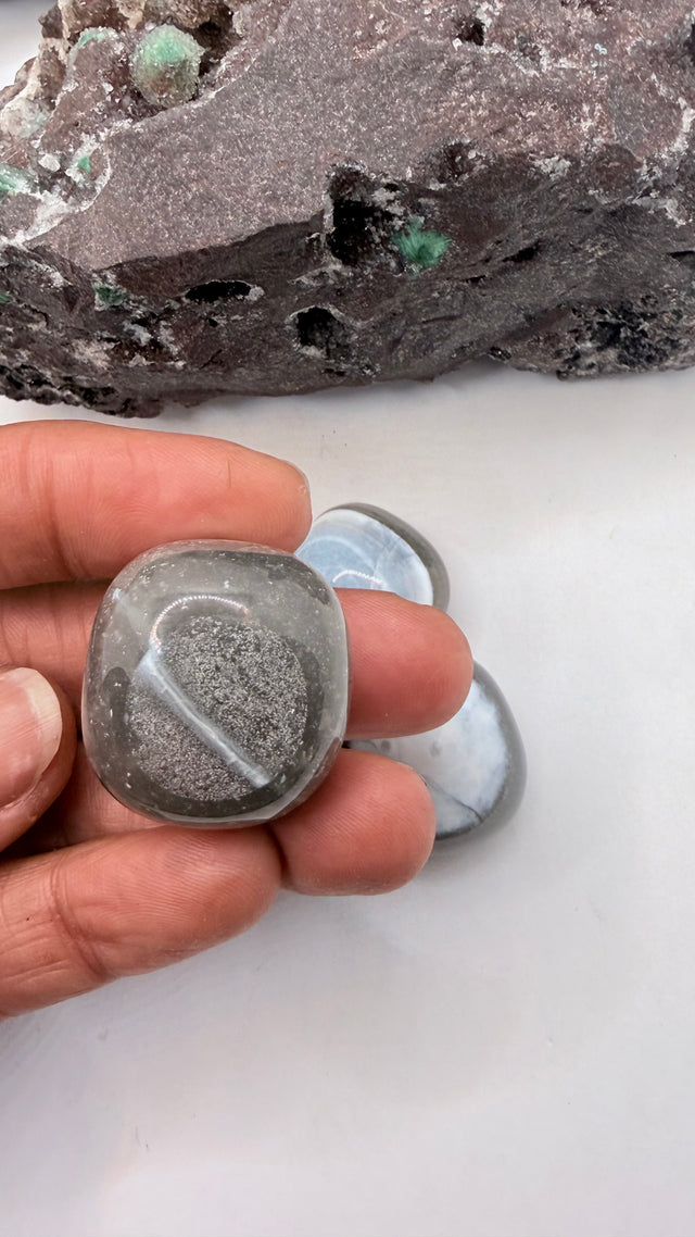 A hand holds a polished gray blue indian opal evil eye 
tumbled stone with a heart-shaped cutout, with two more polished stones and a rough stone in the background on a white surface.